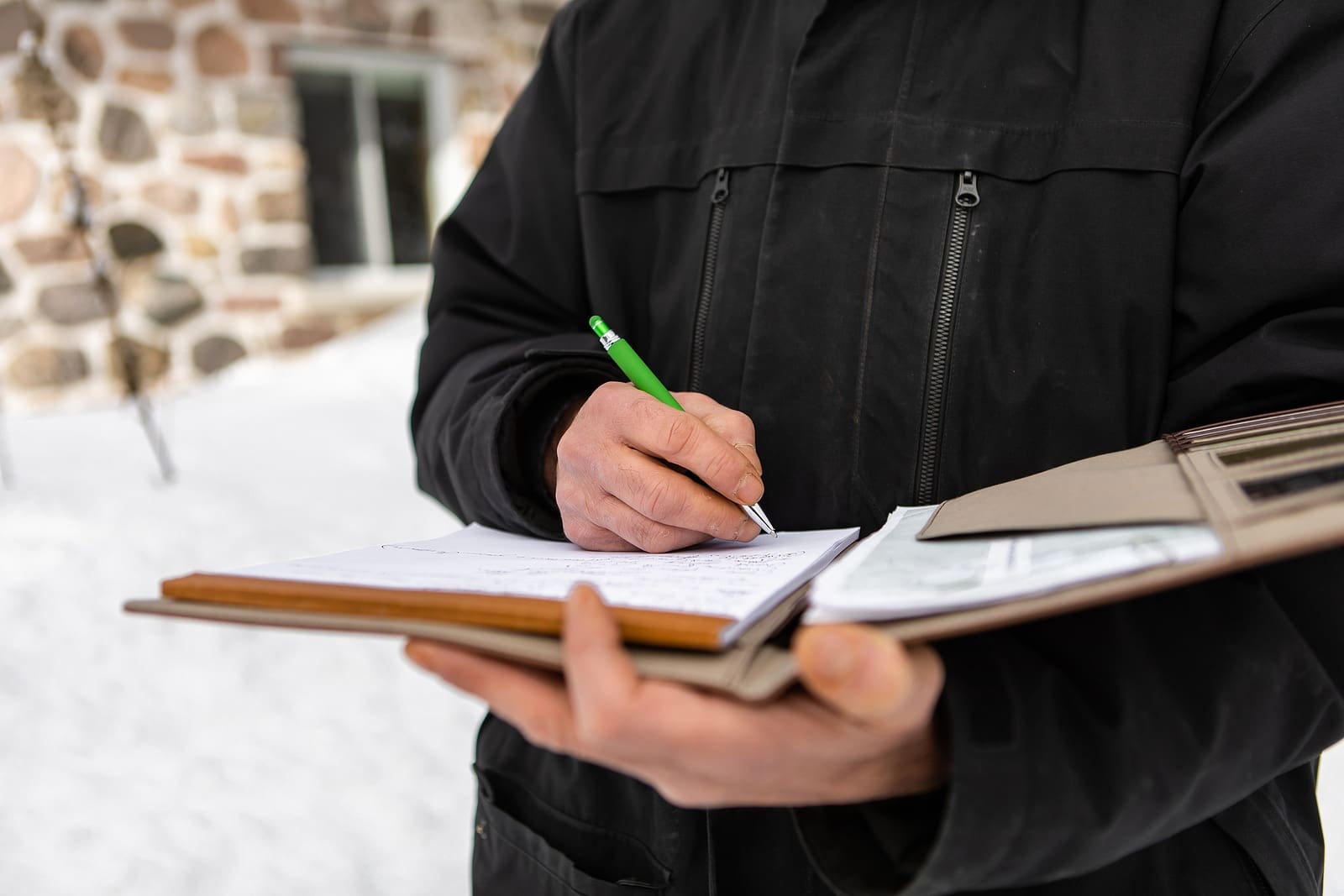 selective focus and close up view of mans hands as he holds a notebook and writes notes, home inspector wearing winter clothes outdoor in snow